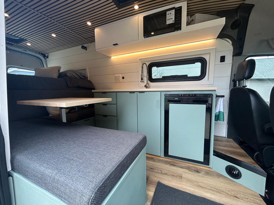 Modern RV rental interior features a mint green kitchenette with a sink, microwave, and fridge, adjacent to a bed and dining area.
