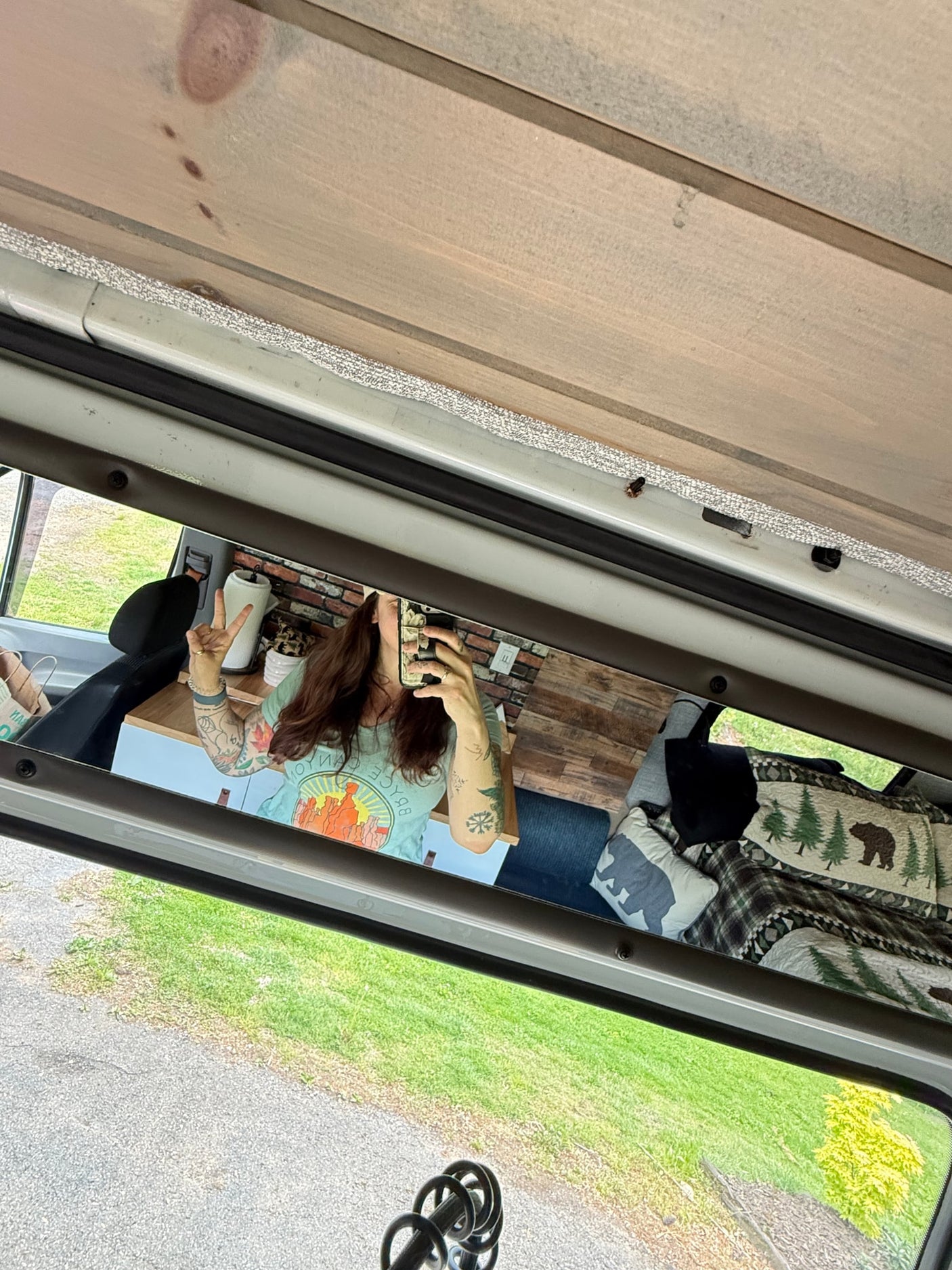 Inside an RV rental, a woman takes a mirror selfie showing off her custom camper interior with wood panels and nature bedding.