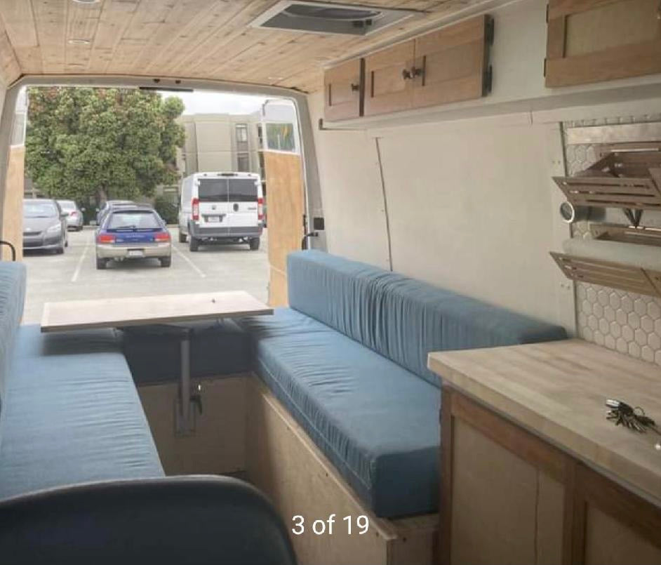 Inside an RV rental: a custom camper van interior with comfy blue seating, a dining table, and wooden cabinets for travel.