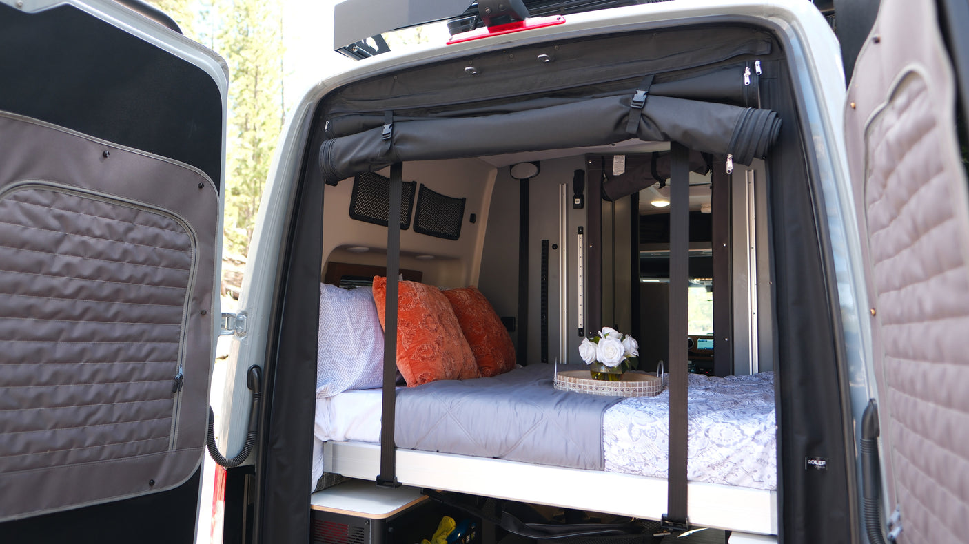 Cozy RV rental bed in a camper van with open rear doors revealing nature. Perfect for van life travel.