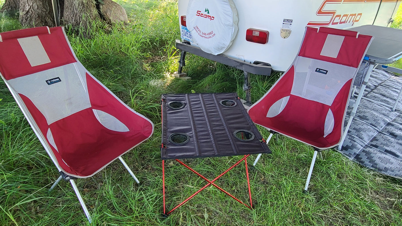 Two red camping chairs and a table are set up on grass, next to a white Scamp RV rental.