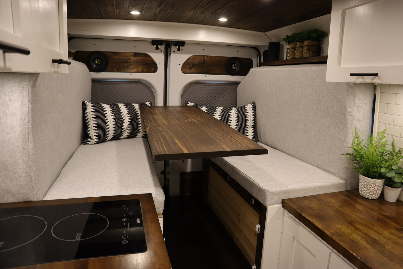 Spacious RV rental camper van interior. Features light grey benches, a wooden table, kitchen counter with cooktop, and storage.