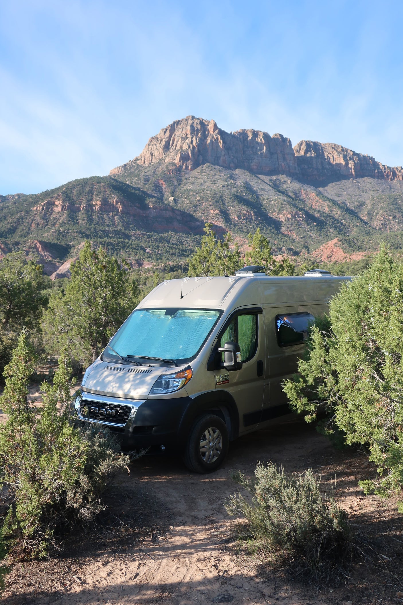 Beige RV rental camper van nestled among trees, with majestic mountains under a blue sky.
