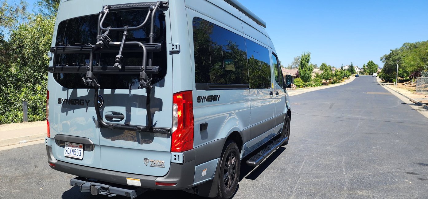Light blue Thor Synergy RV van with bike rack, parked on a residential street. Great option for rv rental.