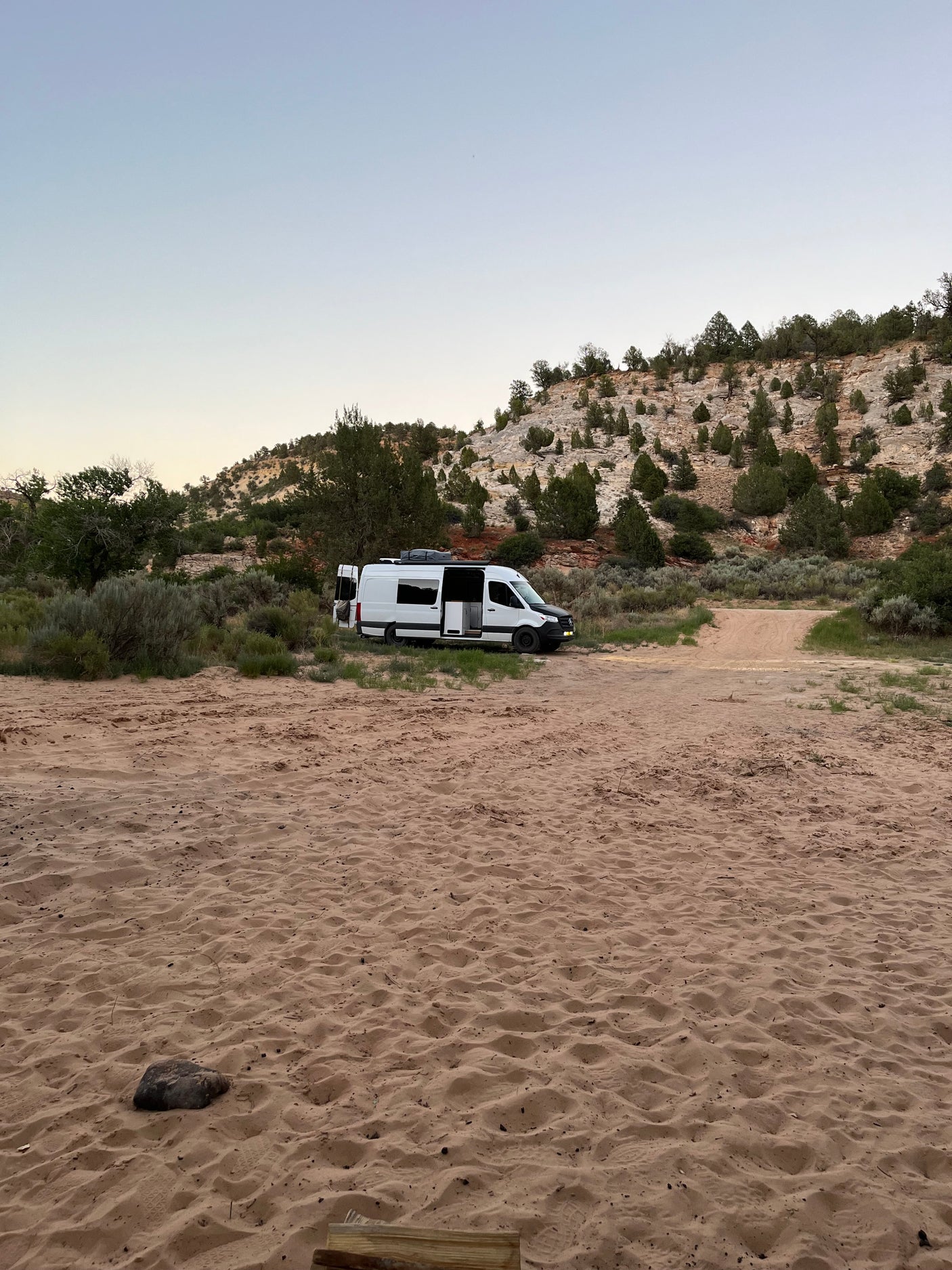 An RV rental van parked in a sandy desert landscape at dusk/dawn with hills and sparse trees.