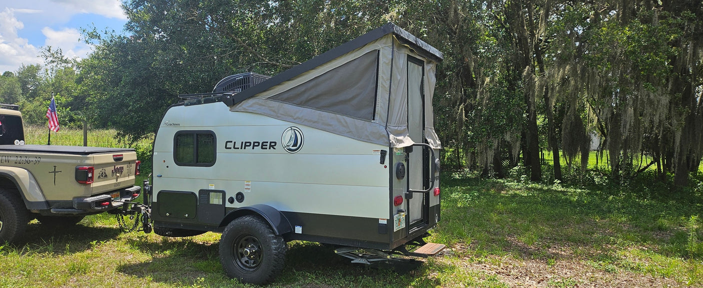 A tan Jeep Gladiator towing a light-colored Clipper pop-up RV rental camper, parked in a green, mossy wooded setting.