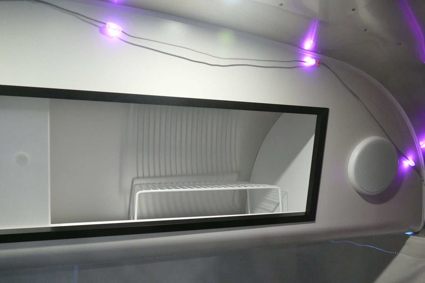 Modern RV rental interior: a clean, white storage unit (likely a refrigerator) with a shelf, illuminated by purple LED lights, and a speaker nearby.