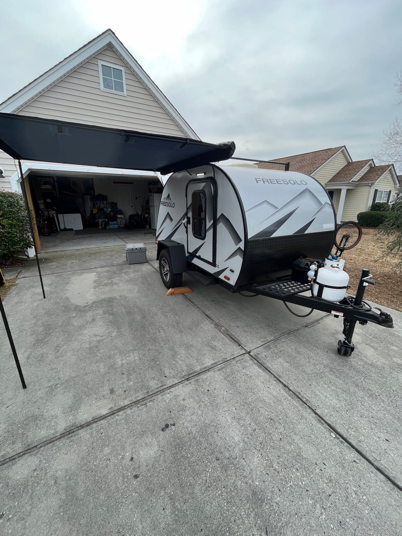 A FREESOLO teardrop RV rental, with an extended awning and propane tank, rests on a driveway beside a house.