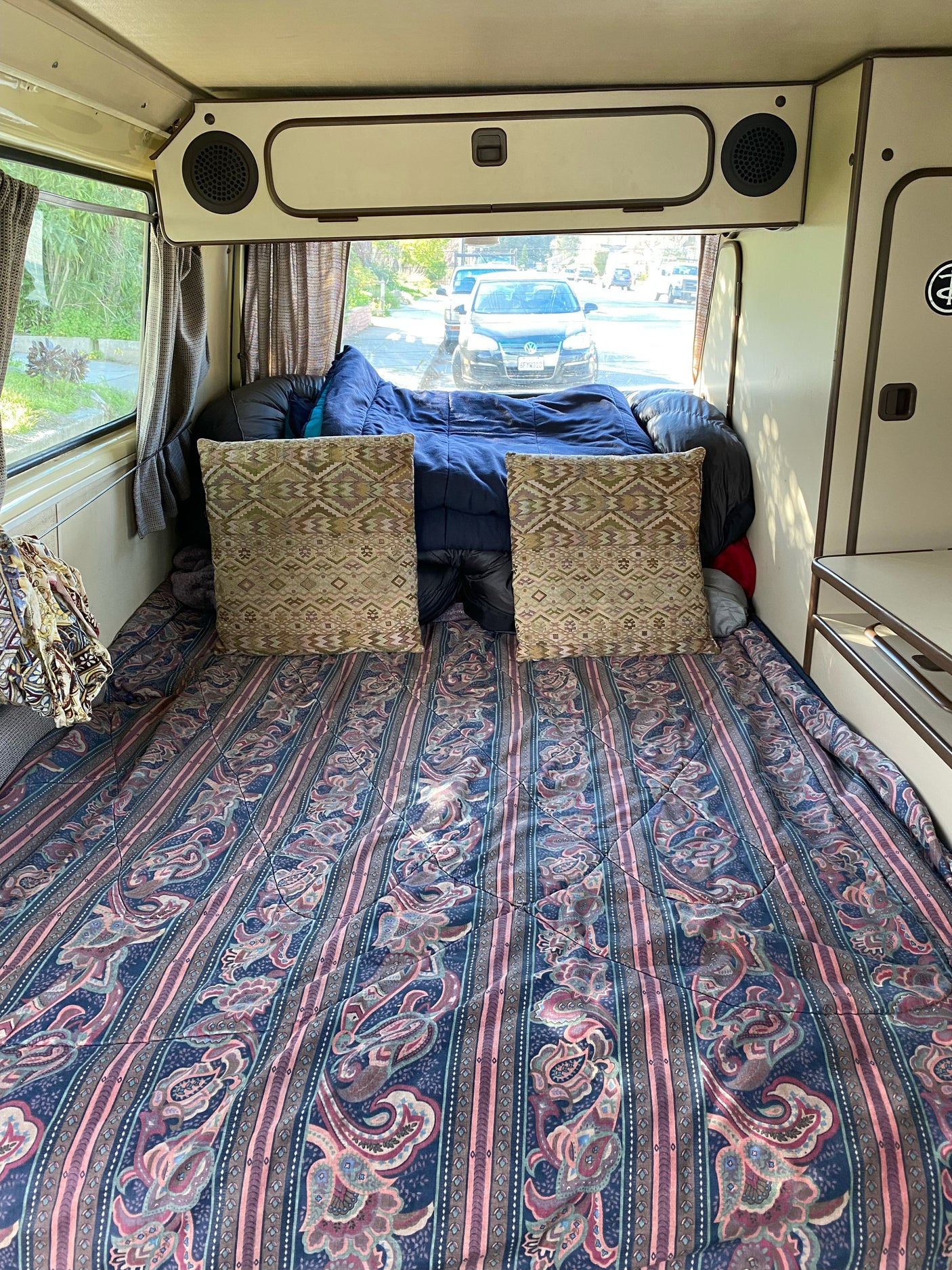Cozy RV rental interior with a comfortable bed, patterned bedding, and a view of the street through the rear window.