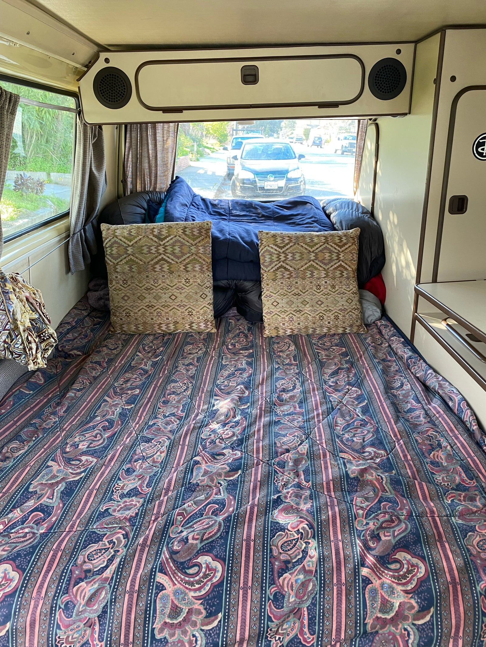 Cozy RV rental interior with a comfortable bed, patterned bedding, and a view of the street through the rear window.