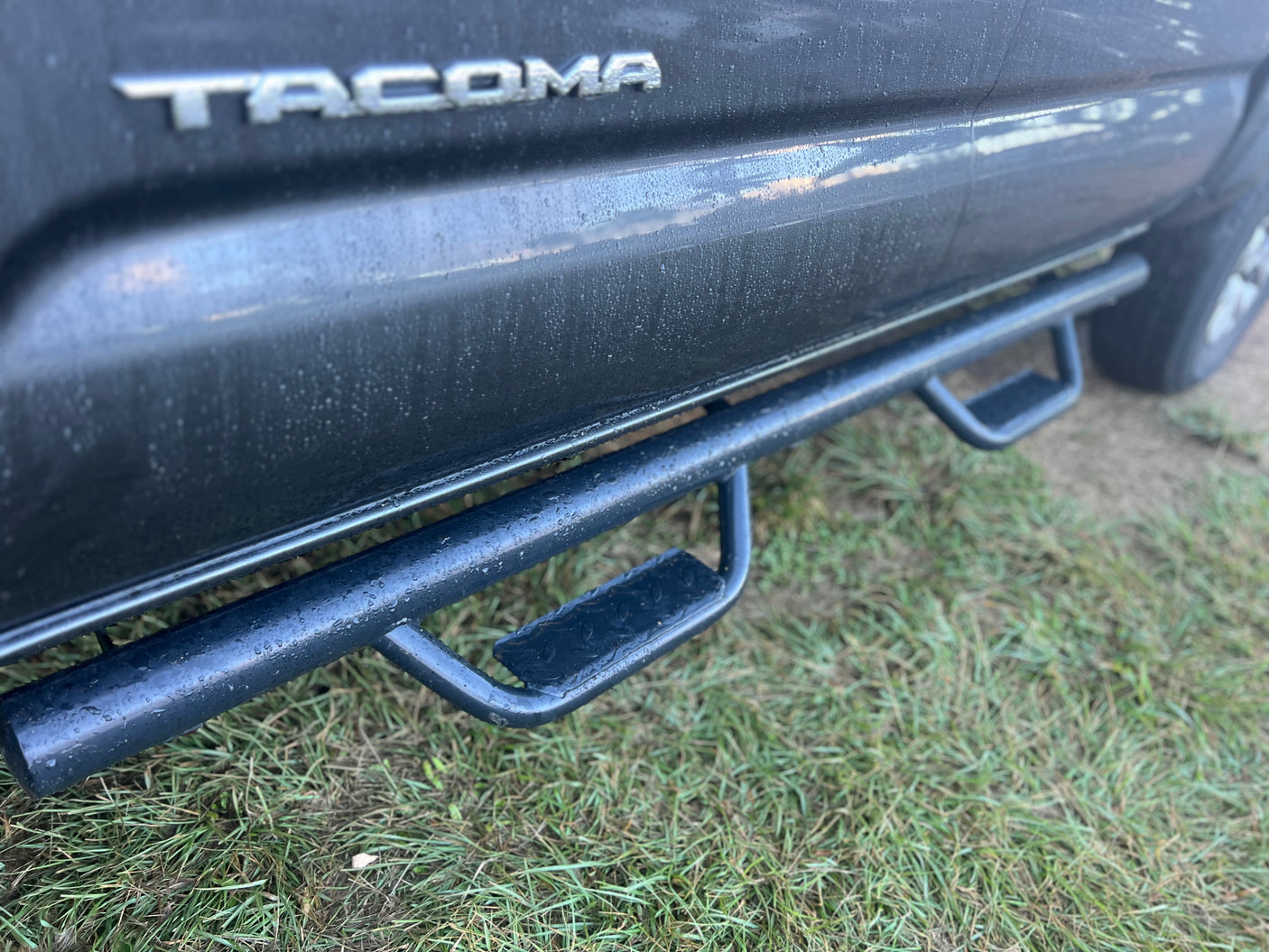 Wet Toyota Tacoma truck, dark paint & sturdy steps on grass. Ready for rugged use alongside your RV rental adventure!