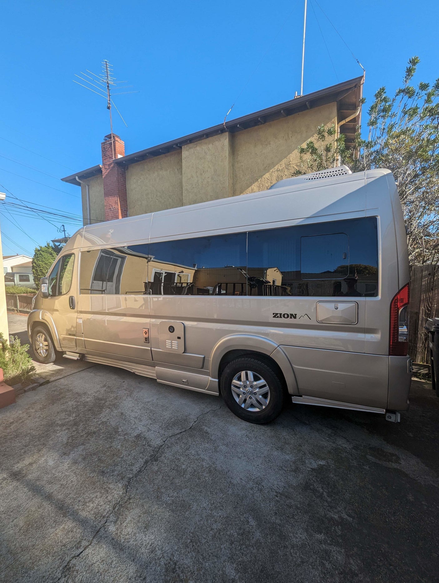 Champagne colored Roadtrek Zion camper van for RV rental, parked on a sunny driveway beside a house.