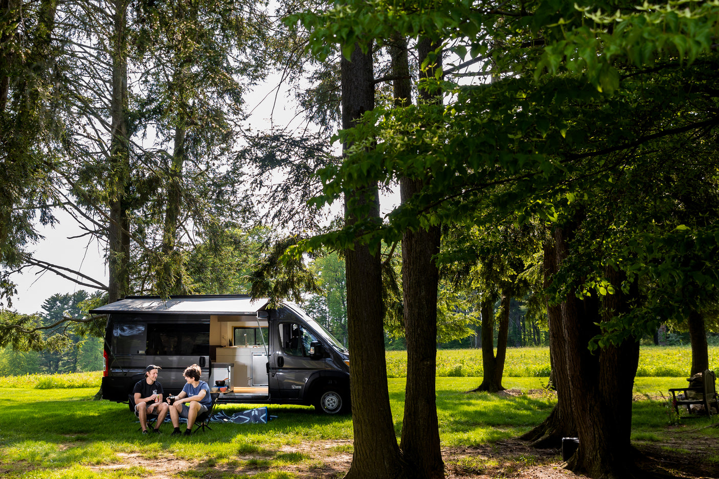 Two friends relax outside their rv rental camper van, parked under shady trees in a bright, grassy field.