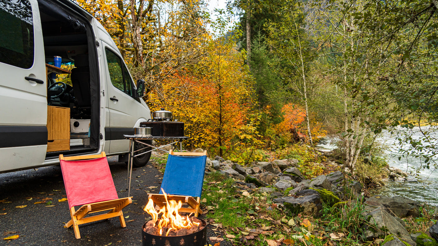 Fall RV rental camping: A camper van beside a vibrant autumn river, with a crackling fire and cozy chairs.