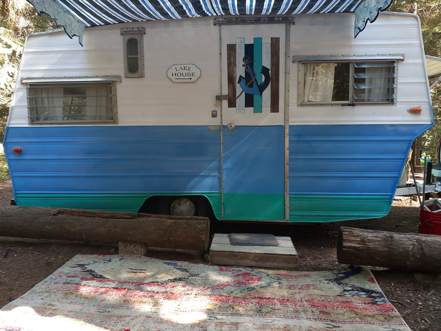 Cozy vintage RV rental, painted blue & teal with anchor door, sign reads "LAKE HOUSE," & outdoor rug.