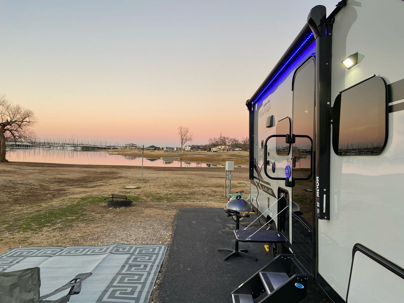 RV rental by a serene lake at sunset. Features a lit travel trailer, outdoor mat, grill, and boats in the tranquil water.