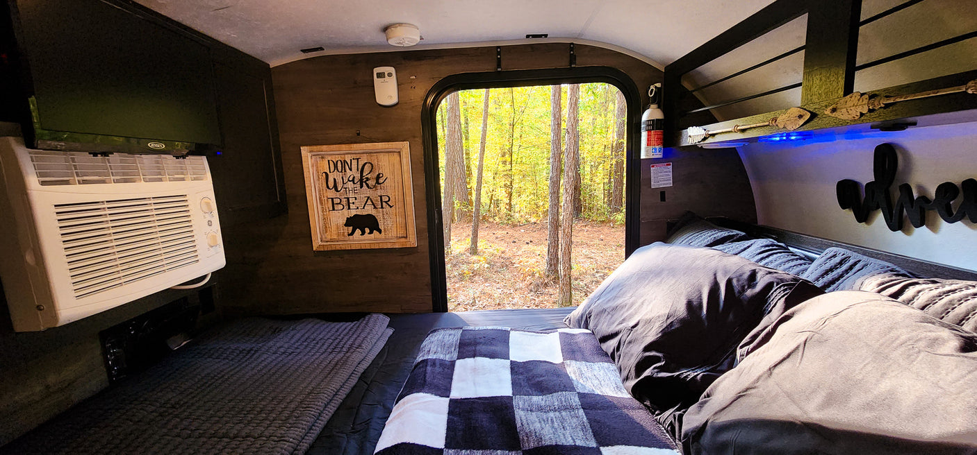 Cozy RV rental bedroom with a forest view, featuring an AC unit, TV, and "Don't Wake the Bear" sign. Perfect for a nature retreat.