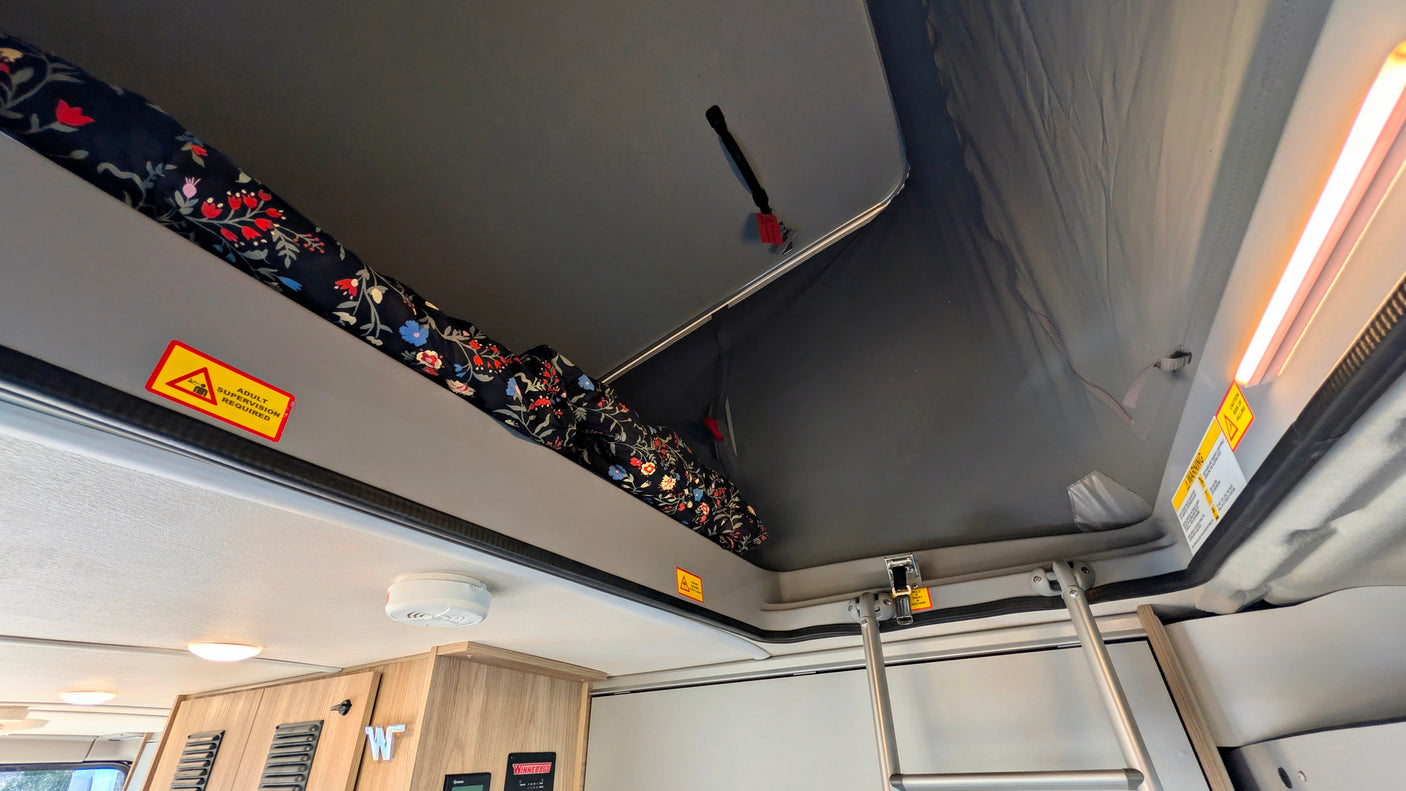 Elevated sleeping area inside an RV rental with pop-top roof, floral bedding, safety warnings & ladder.