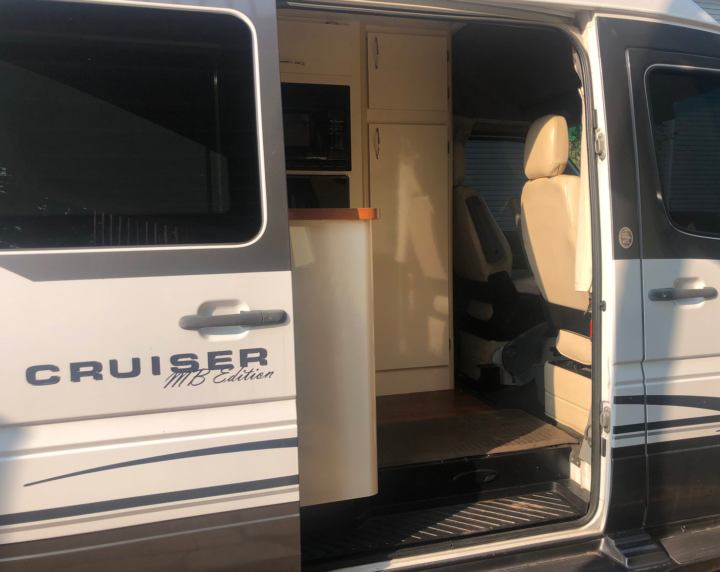 "CRUISER MB Edition" rv rental: Open side door reveals van interior with compact kitchen (microwave, cabinets) and light seating.