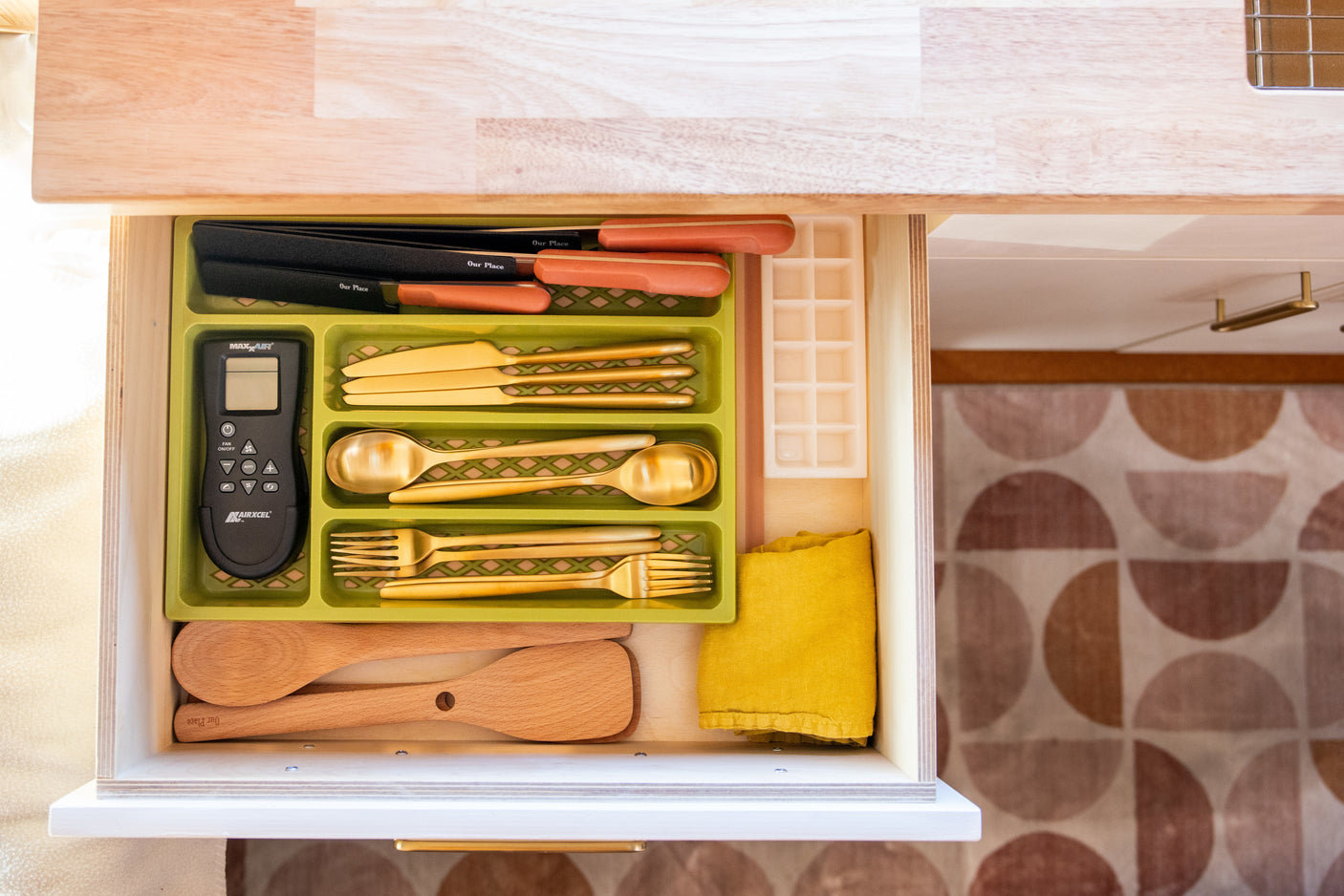 Organized RV rental kitchen drawer with golden cutlery, MaxxAir remote, Our Place knives, and wooden utensils.