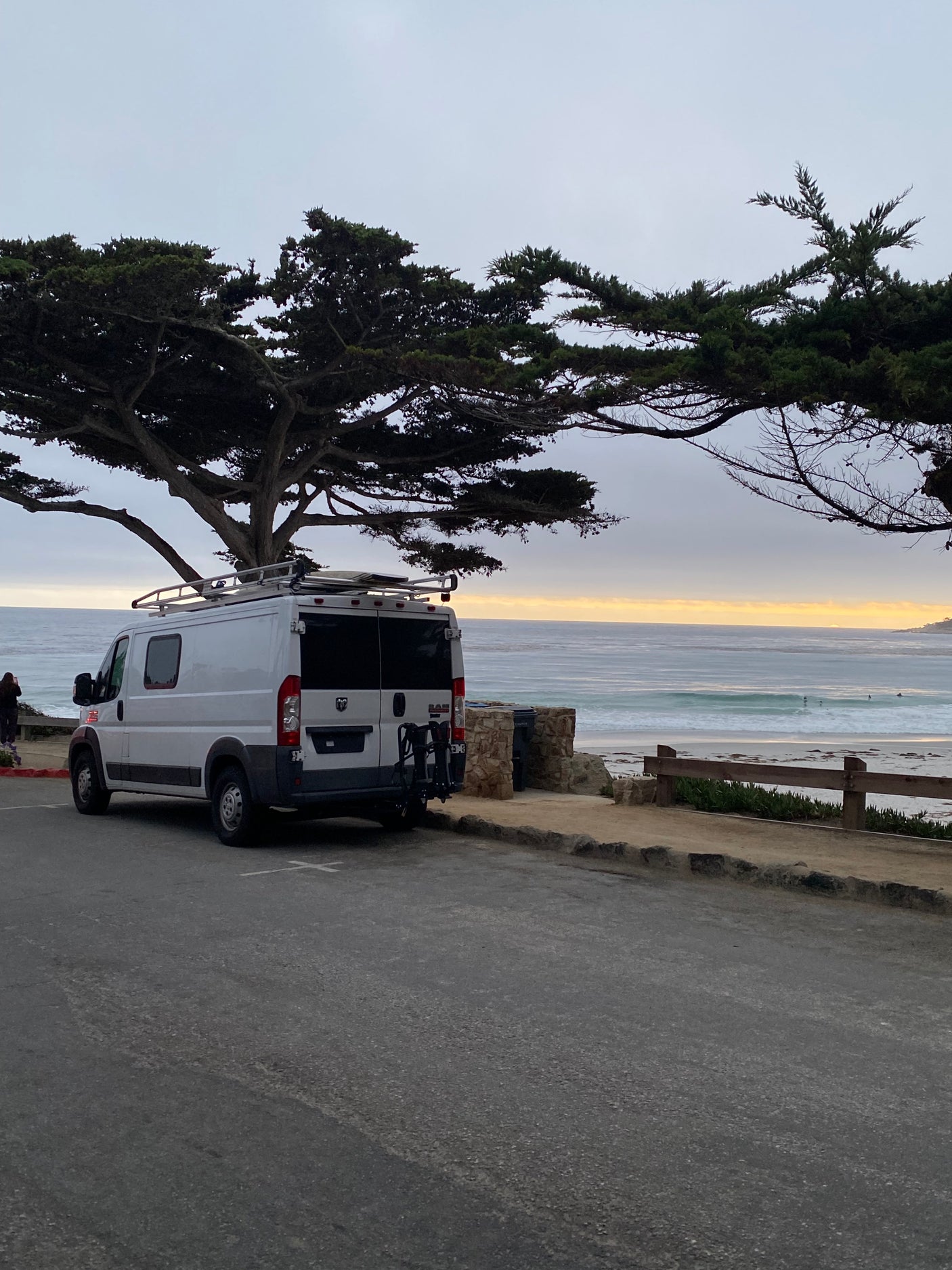 White RV rental parked beachfront under a large tree, overlooking ocean waves, with surfers and a golden horizon.