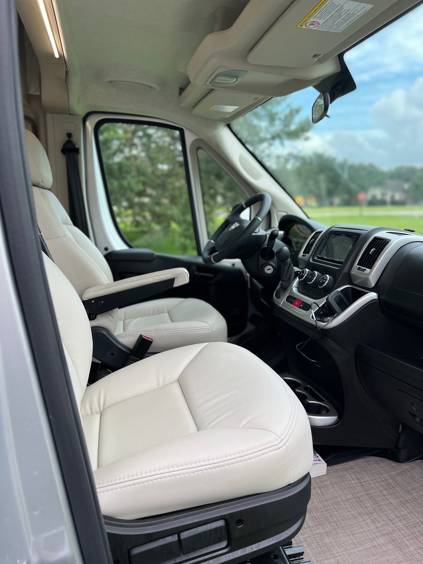 RV rental's front cabin. Cream-colored seats with armrests, steering wheel, and dashboard with infotainment screen.