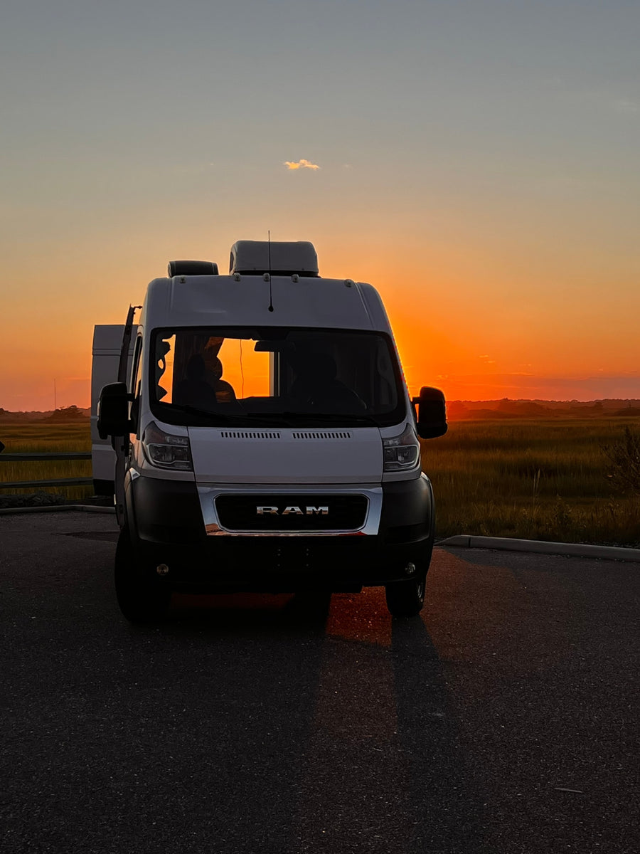 A white Ram ProMaster camper van, an ideal RV rental, sits on asphalt, silhouetted by a vibrant orange sunset over marshlands.