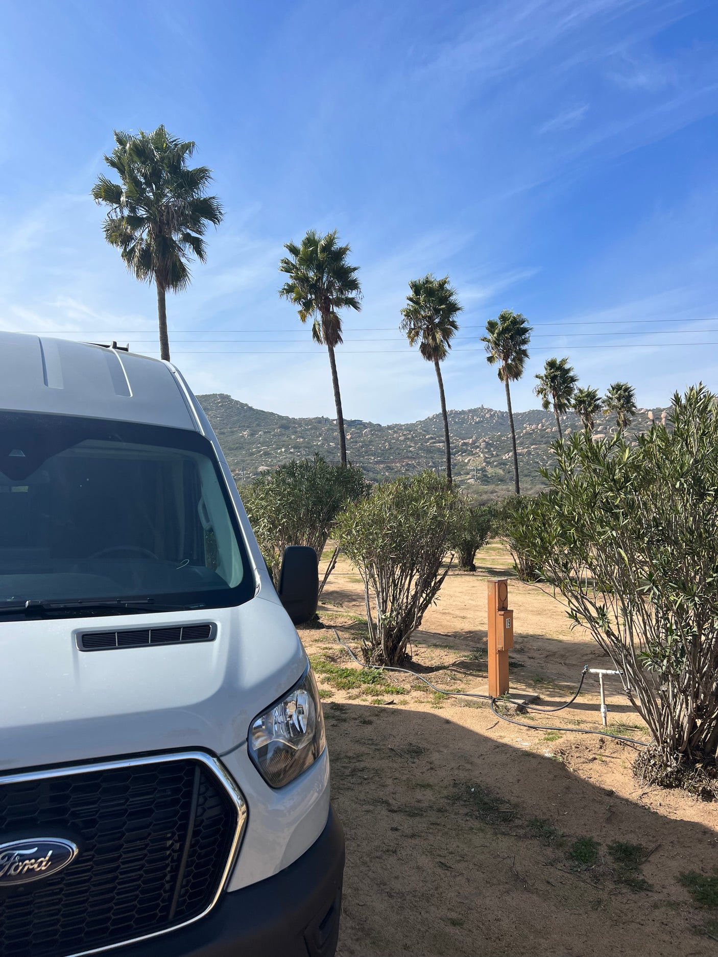 White Ford Transit RV rental van parked at a sunny campsite with utility hookups, palm trees, and distant hills.