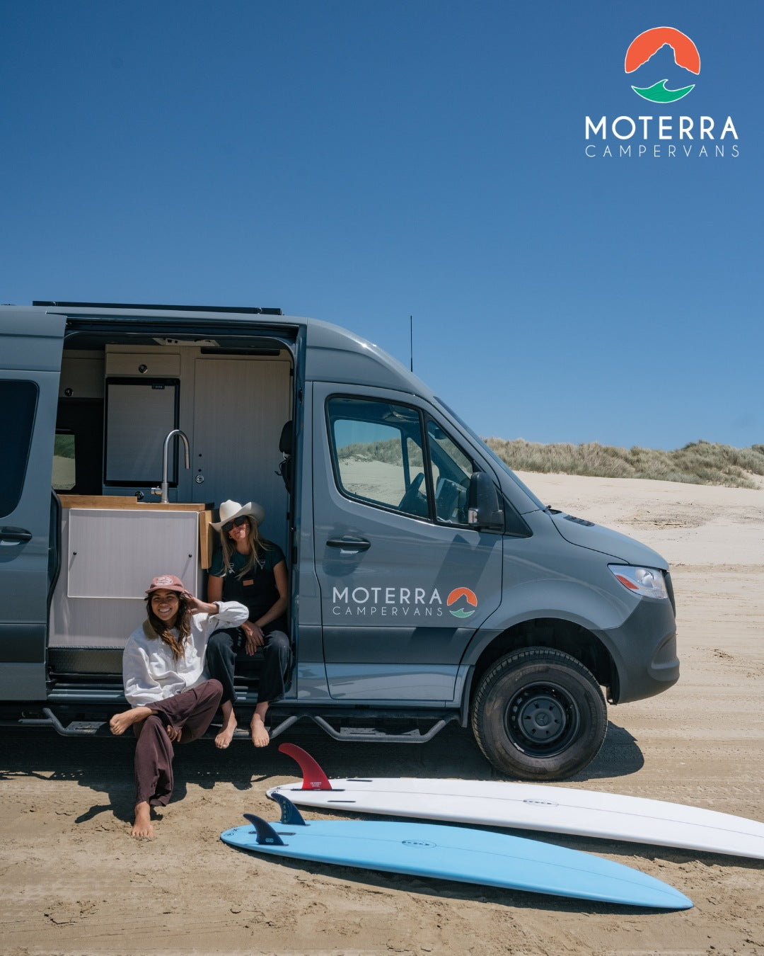 Enjoy the freedom of an RV rental! Two women relax with surfboards by their Moterra campervan on a sunny beach.