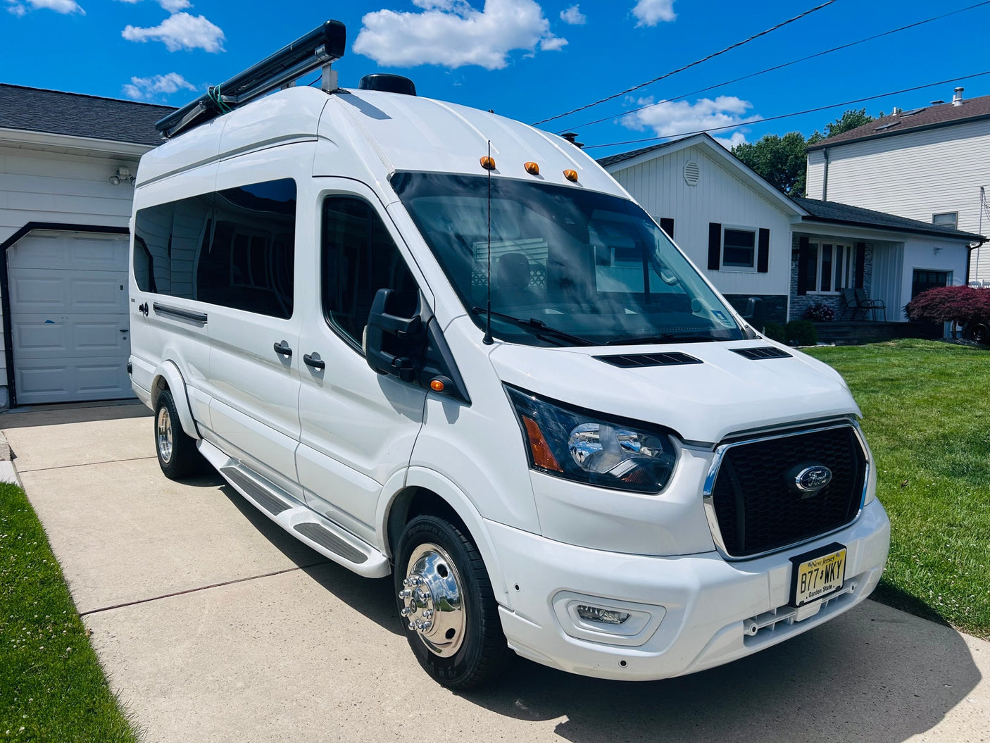 White Ford Transit camper van with roof awning and dually wheels, parked on a sunny driveway, ready for RV rental.