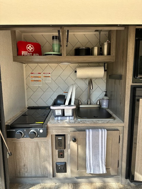 Inside an RV rental: compact kitchen featuring a sink, stove, microwave, and neatly organized cooking essentials.