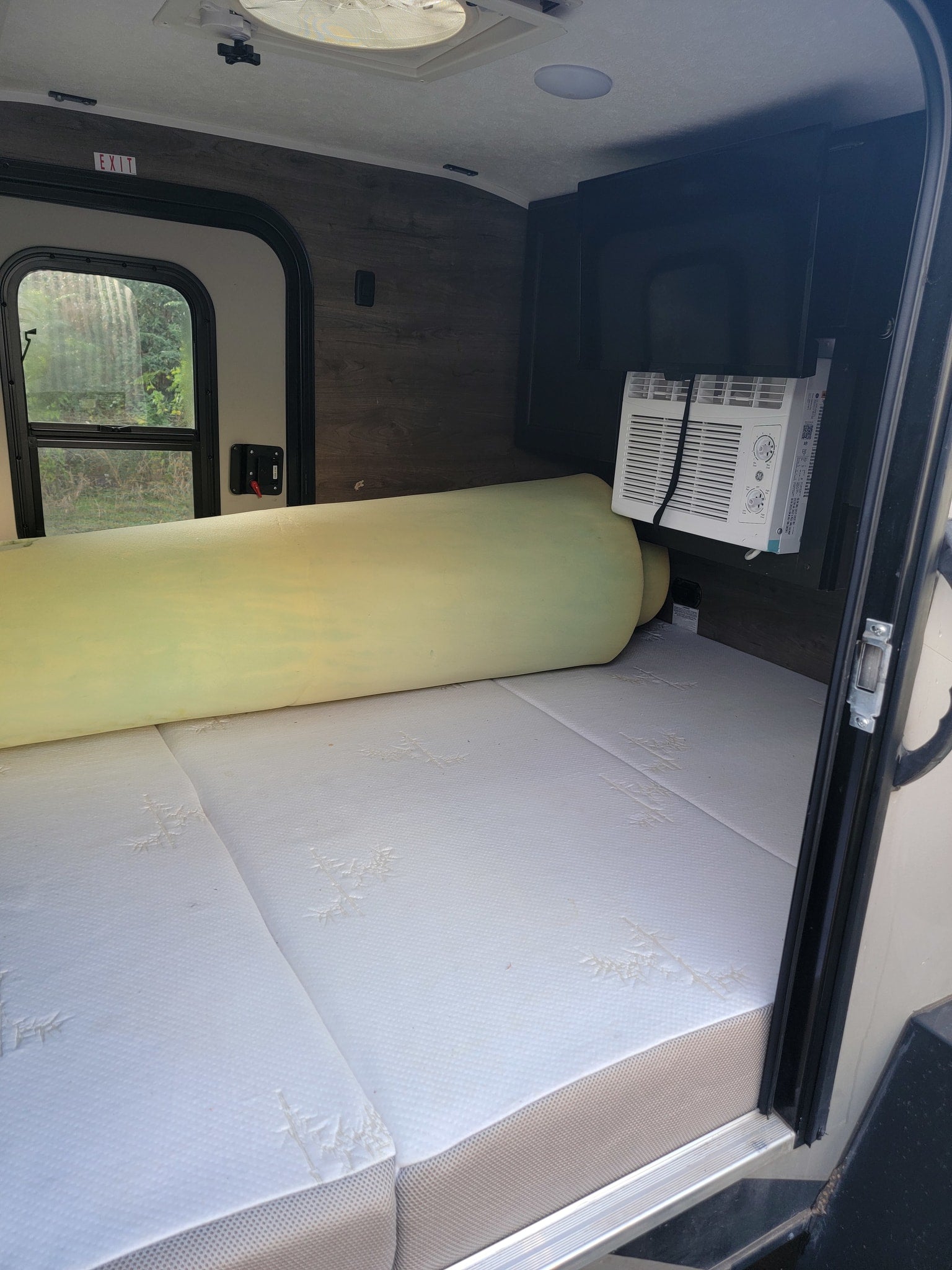 RV rental interior: A mattress with a rolled foam topper, a window AC unit, and an EXIT sign above the side window.