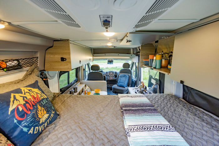 and open back doors reveal a serene view of a stream flowing through lush green trees.|Cozy camper van interior with elevated bed