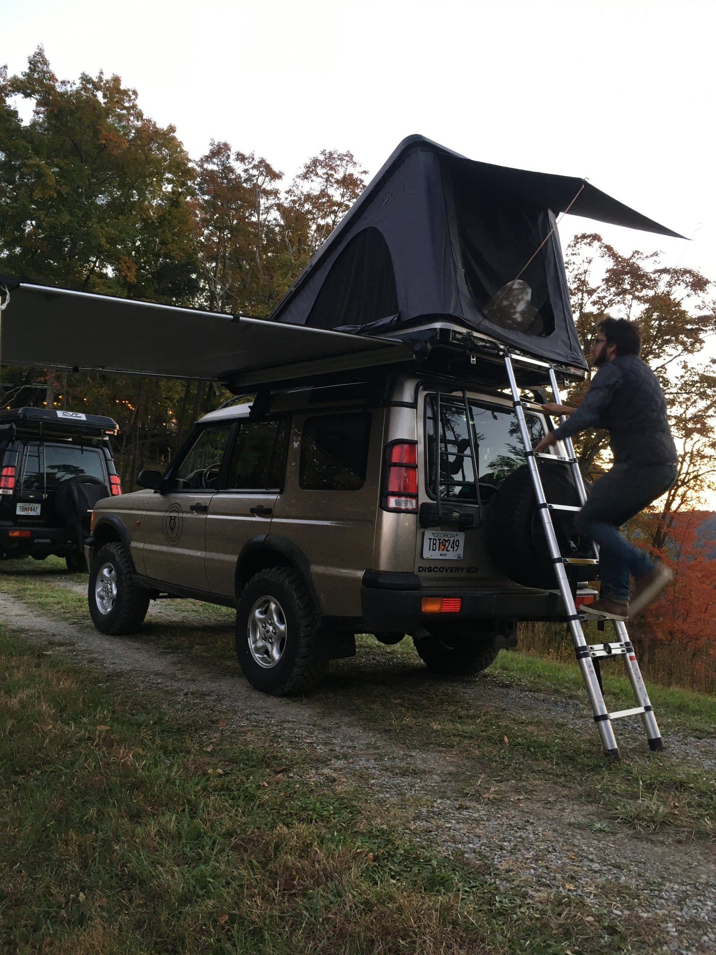 A person climbs a ladder to a rooftop tent on a Land Rover Discovery.