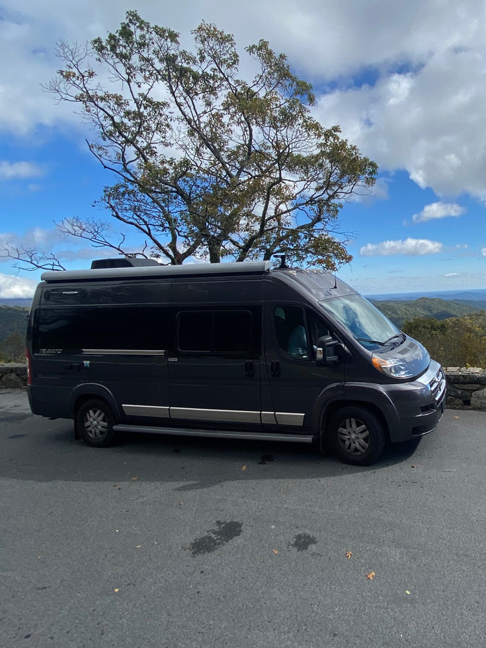 offering stunning views of valleys & autumn foliage under a blue sky.|Experience freedom with an RV rental! This dark gray camper van enjoys a stunning mountain overlook view under a beautiful sky.|Black RV rental parked in an autumn campsite with extended awning