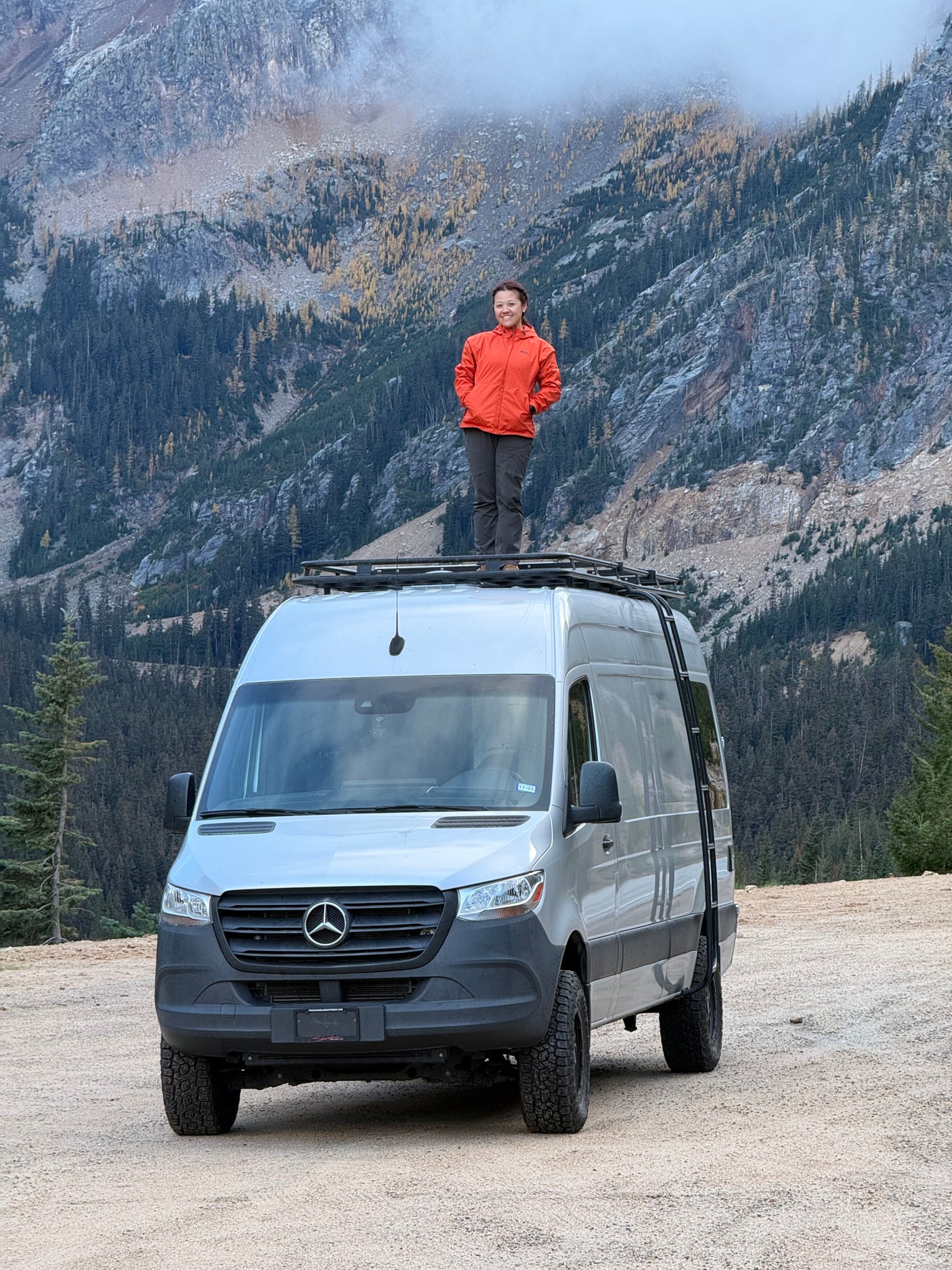 A woman enjoys mountain views from her Mercedes Sprinter RV rental rooftop, with misty peaks and autumn larches in the background.