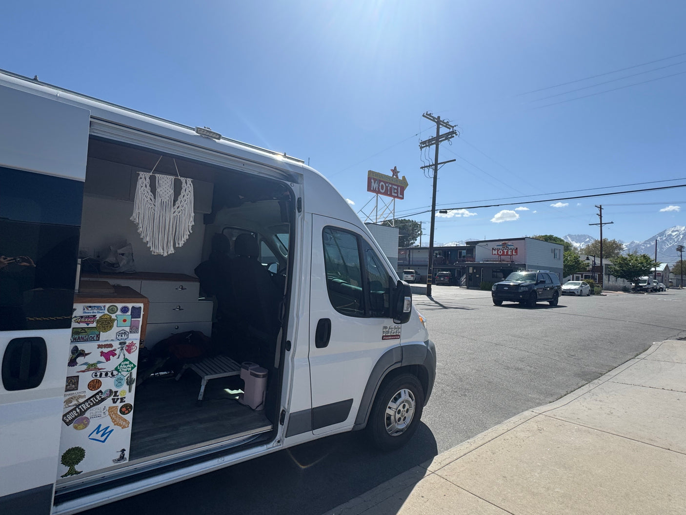 A white camper van, likely an RV rental, with its side door open, parked by a motel and distant mountains on a sunny day.