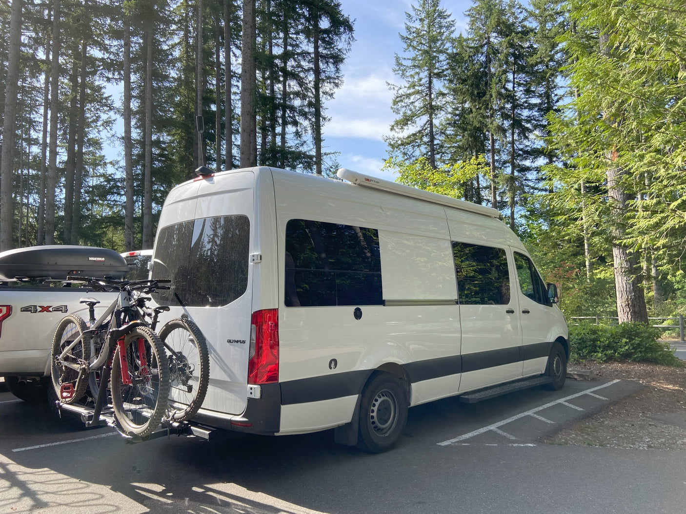 An RV rental camper van with mountain bikes on a rack, parked in a forest setting on a sunny day.