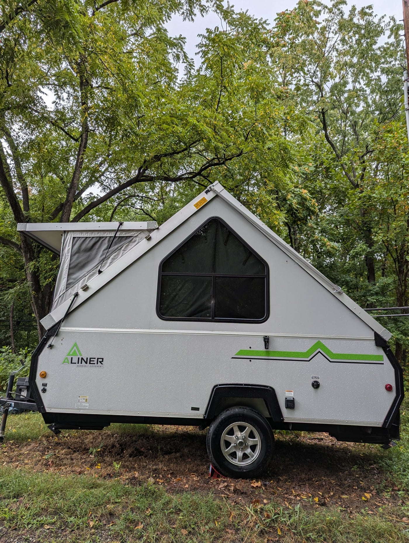 A gray A-liner A-frame pop-up camper, suitable for rv rental, parked in a lush, tree-filled setting with rain droplets visible. — photo 2