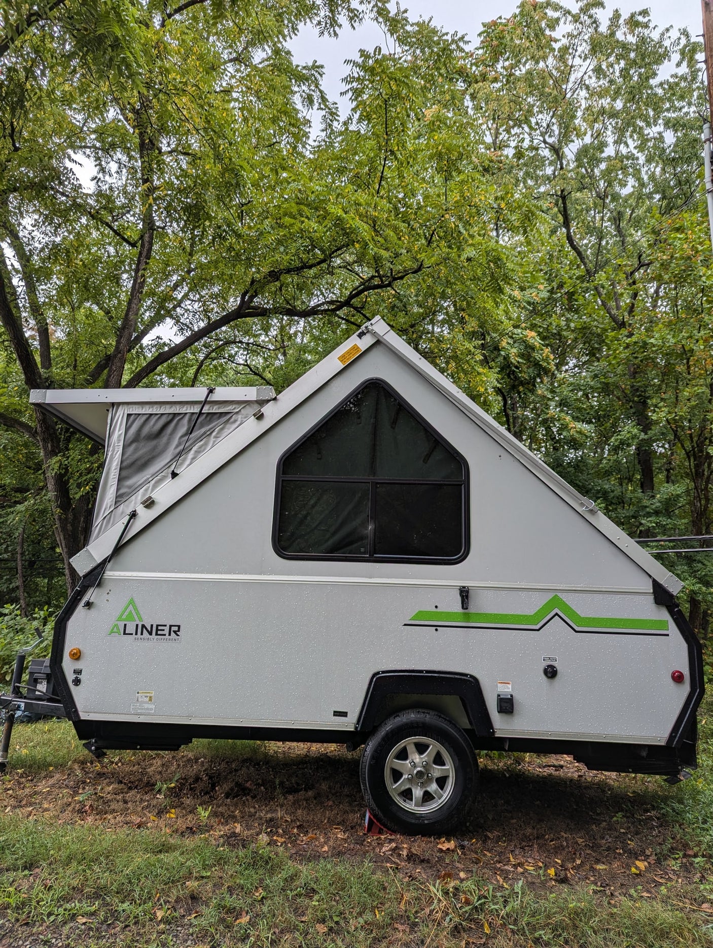A gray A-liner A-frame pop-up camper, suitable for rv rental, parked in a lush, tree-filled setting with rain droplets visible.
