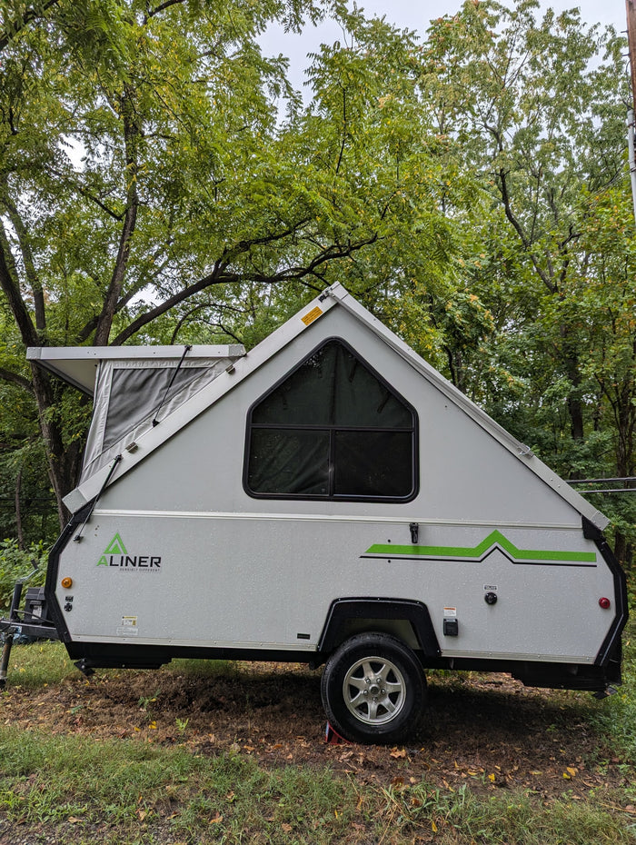 A gray A-liner A-frame pop-up camper, suitable for rv rental, parked in a lush, tree-filled setting with rain droplets visible.