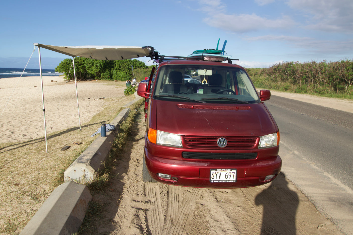 Maroon VW van, an RV rental, parked on a sandy spot next to a Hawaii beach. Awning extended, surfboards on roof, ready for camping.