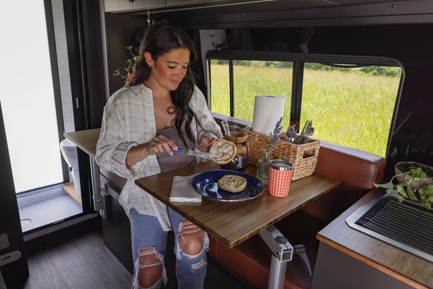 Enjoy breakfast in an RV rental. A woman spreads topping on an English muffin at a table with coffee, overlooking a field.