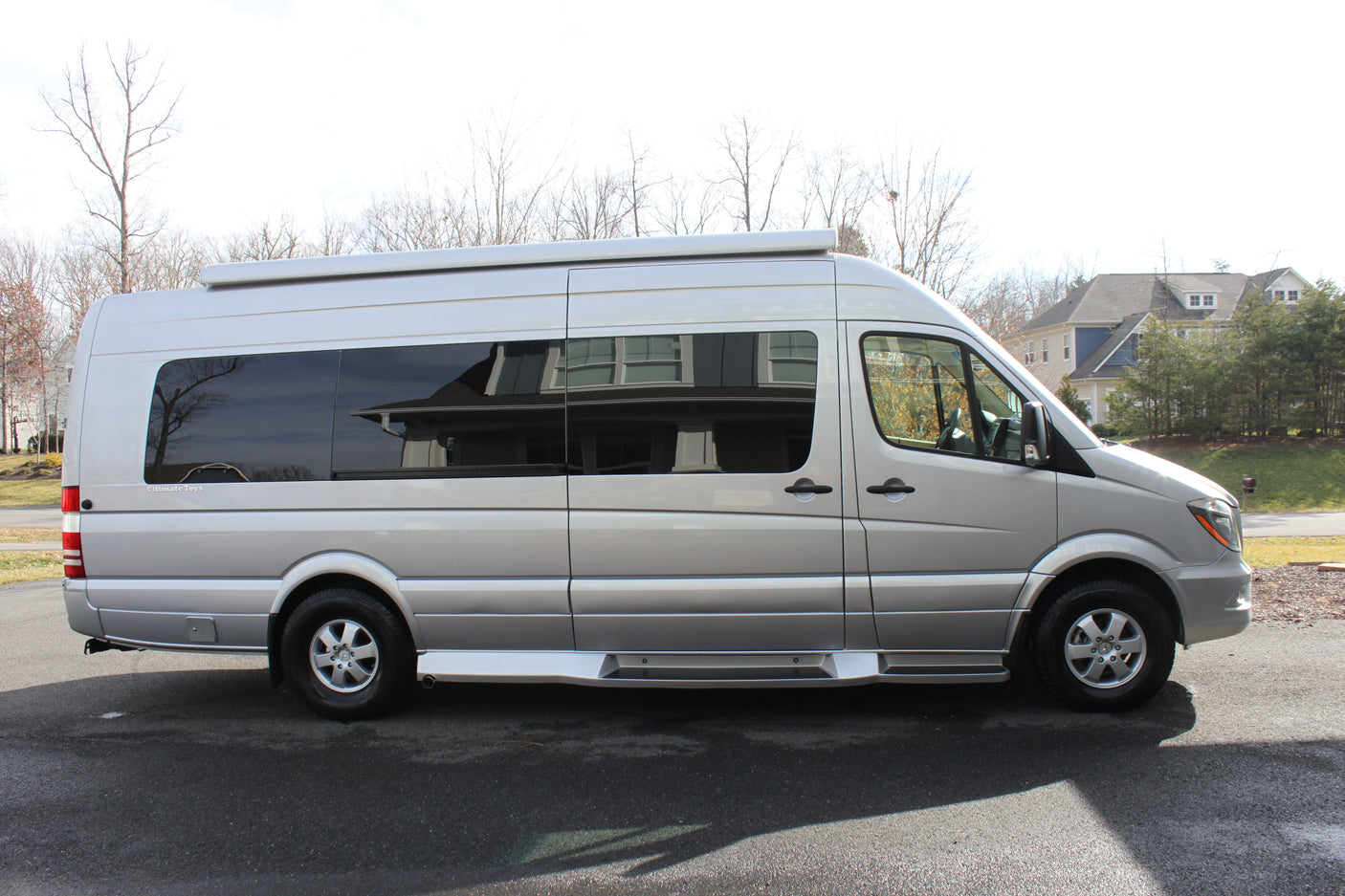 A silver Mercedes-Benz Sprinter camper van, ideal for RV rental. Seen from the side, parked on asphalt with houses and trees.