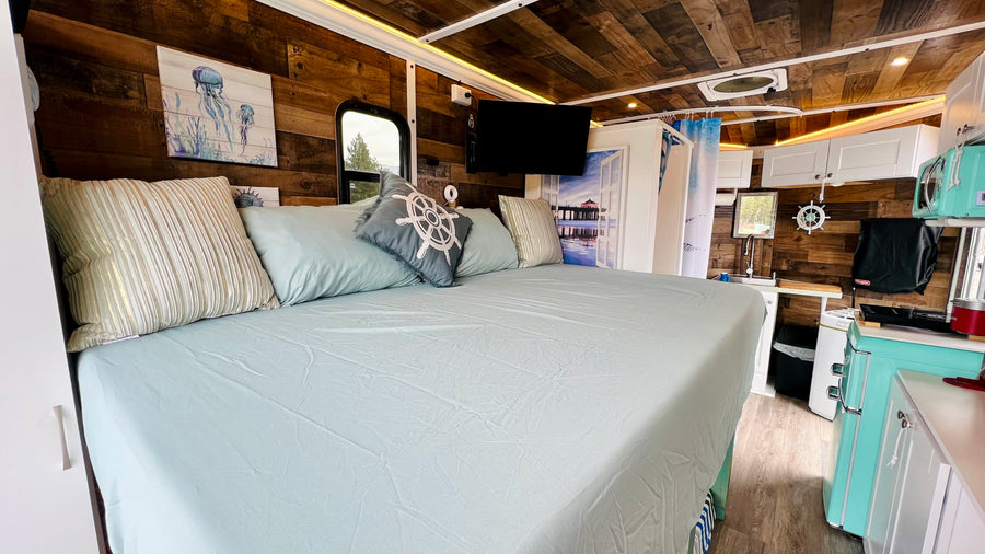 Spacious RV rental interior featuring a large bed, nautical decor, compact kitchen with retro fridge, and shower.