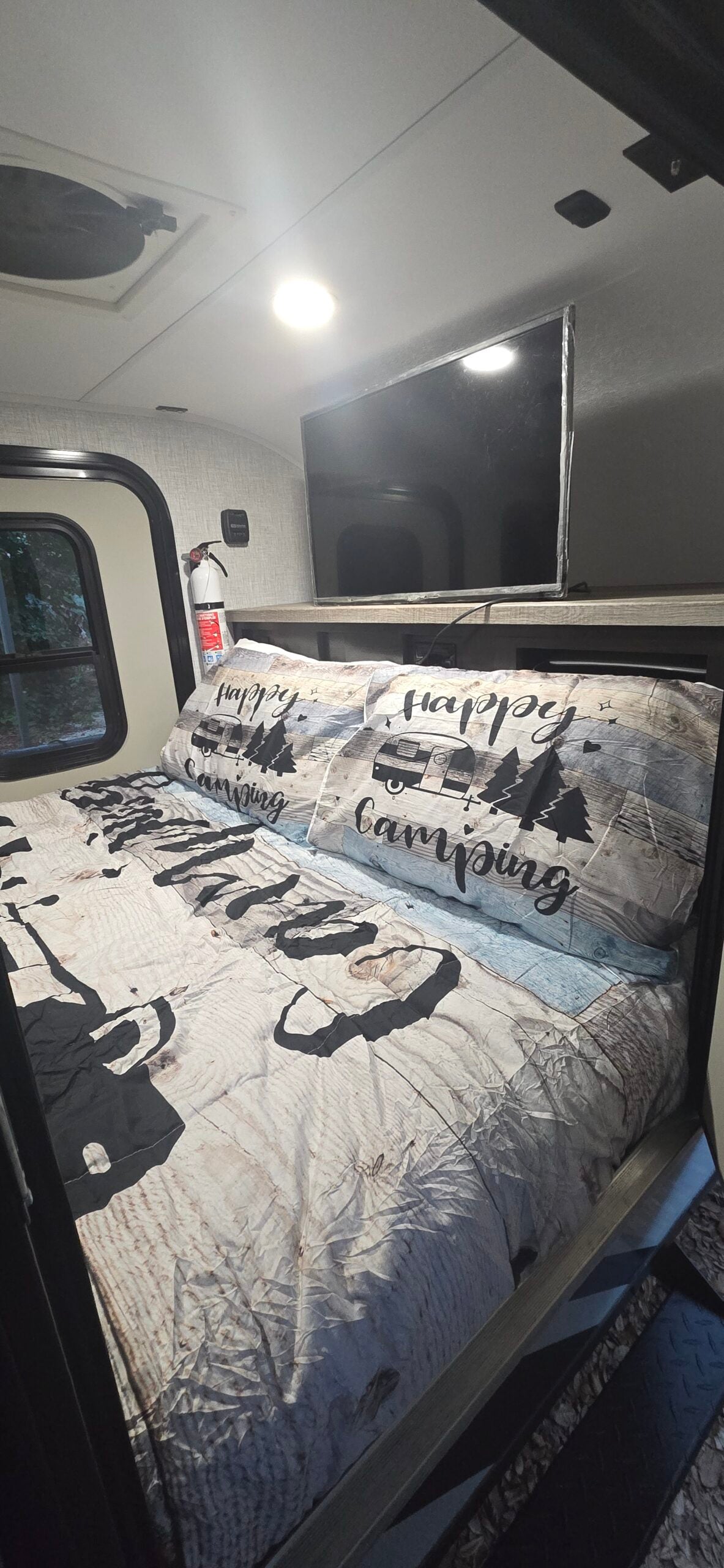 Cozy RV rental bedroom featuring 'Happy Camping' themed bedding, a TV, and a window offering a view of outdoor greenery.