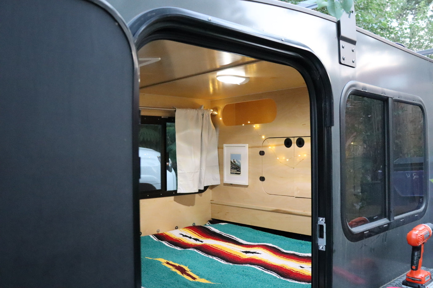 Cozy RV rental interior shows a colorful bed, wooden walls, and string lights, viewed from the open doorway.