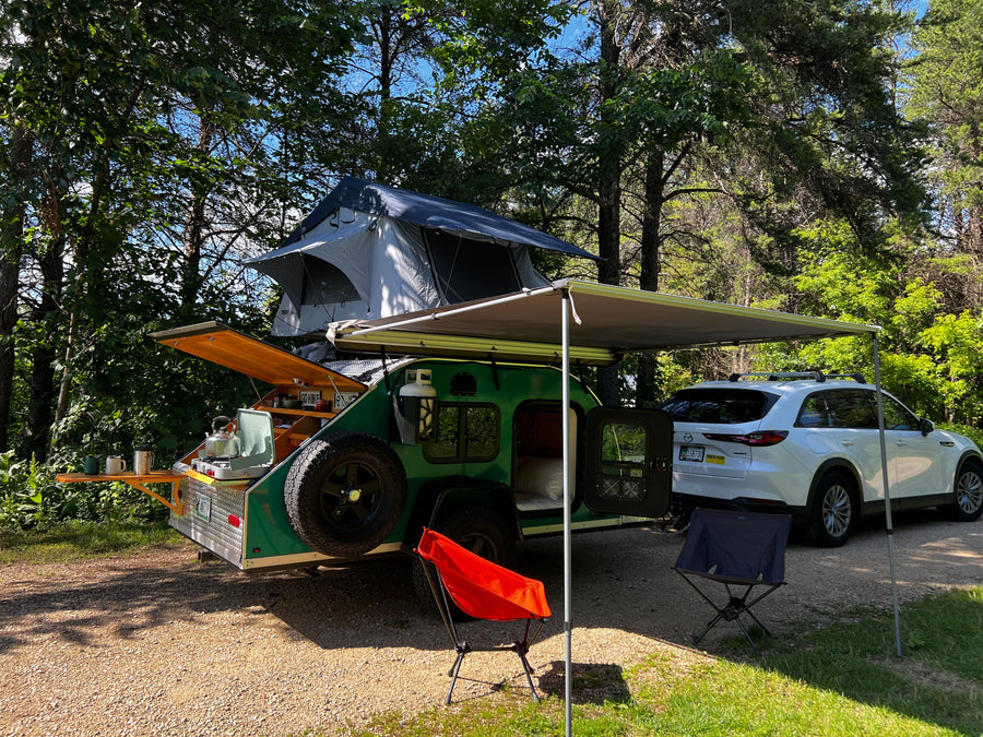 Enjoy nature with this RV rental! A green teardrop trailer, rooftop tent, and outdoor kitchen under an awning for a cozy camping trip.