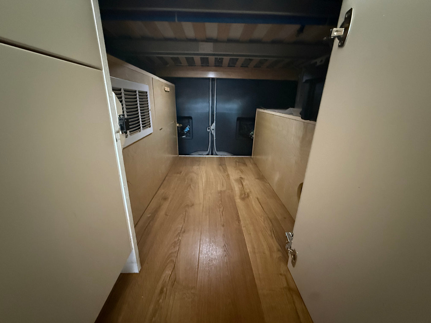 RV rental camper van's under-bed storage: wood-look floor, light panels with vent on left, and rear access area.