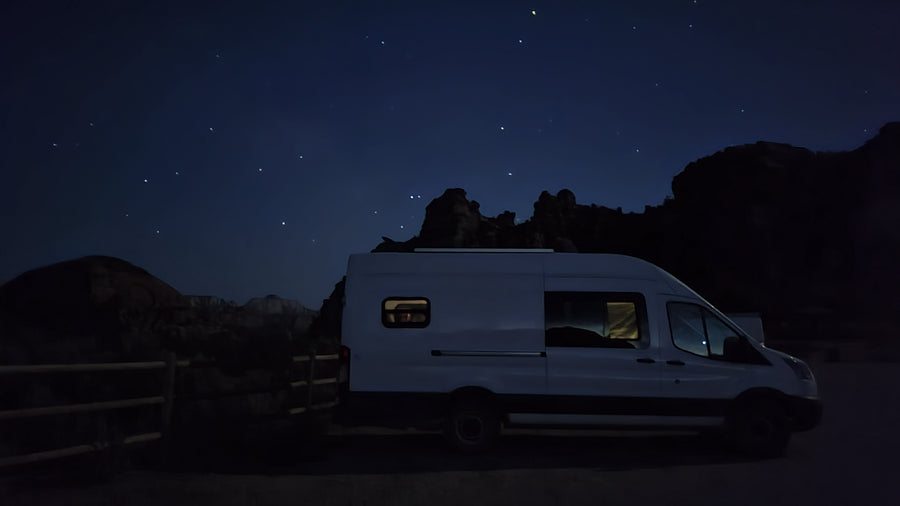 White camper van RV rental parked at night with illuminated interior, under a clear, starry sky & dark rocky terrain.